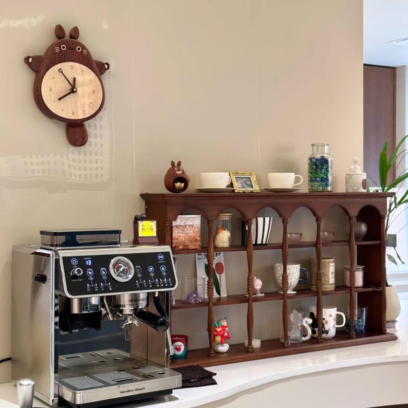 May include: A stainless steel espresso machine with a brown wooden shelf unit behind it. The shelf unit has a variety of decorative items, including a clock, a figurine, and a jar of marbles.