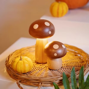 May include: Two wooden mushroom-shaped lamps with white polka dots on their caps sit on a woven tray. A small yellow pumpkin is next to the lamps. The lamps have a warm, inviting glow, suggesting they are turned on.