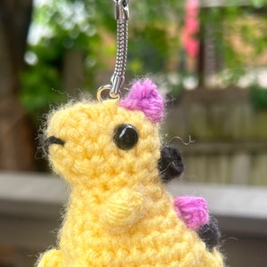 May include: A yellow crocheted dinosaur keychain with purple and black accents. The dinosaur has a small pink bow on its head and black eyes. The keychain is attached to a silver key ring.