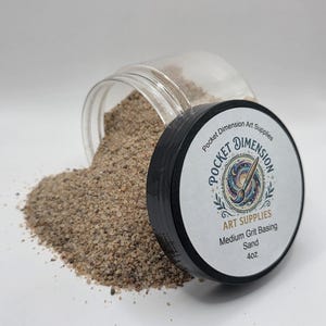 May include: A clear plastic jar with a black lid, partially open, spilling medium grit basing sand. The lid features a label with the text "Pocket Dimension Art Supplies" and "Medium Grit Basing Sand 113g."