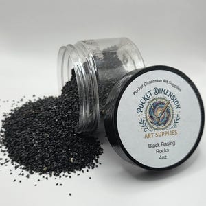 May include: A clear plastic jar, spilling black basing rocks onto a white surface. The jar's black lid, labelled "Pocket Dimension Art Supplies," lies beside it. The label indicates the product contains 113g of black basing rocks.