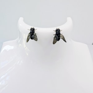 May include: A pair of black fly-shaped earrings. The earrings are displayed on a white mannequin head. The fly earrings have detailed wings and body features, creating a unique and edgy accessory.