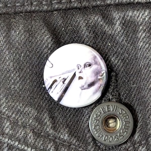 May include: A round, white button with a surreal image of a pale face with an open mouth. The button is next to a silver button with the text "LEVI STRAUSS & CO. CAL. CO." on a black denim jacket.