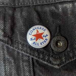 May include: A white button with blue and red lettering that reads "Converse Chuck Taylor All Star". The button has a red star in the center.