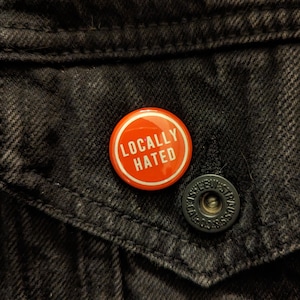 May include: A red and white button with the text "Locally Hated" on a black denim jacket.