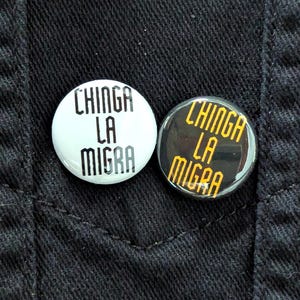 May include: Two round buttons with the text "CHINGA LA MIGRA". One button is white with black text, and the other is black with yellow text. The buttons are on a black fabric background.