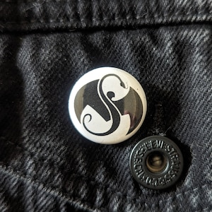 May include: A white button with a black and white logo of a stylized 'S' with wings. The button is on a black denim background.