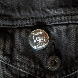 May include: A black and white button with a graphic of a person wearing sunglasses and the text "The Cramps Human Fly".