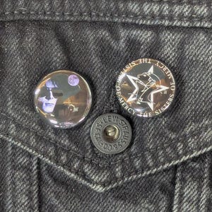 May include: Close-up of a black denim jacket pocket with three metal buttons. Two round pinback buttons are visible, one with a portrait and the other with a star design and the text "Sisters of Mercy".