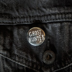 May include: A silver button with the text "Ghost Hunter" in white lettering.