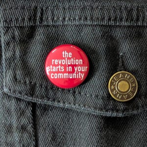 May include: Red button with white text that reads "the revolution starts in your community". The button is pinned to a black denim jacket.