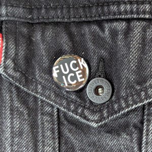 May include: A round, silver-toned button with the words "FUCK ICE" in white block letters. The button is pinned to a black denim jacket near a pocket and a silver button.