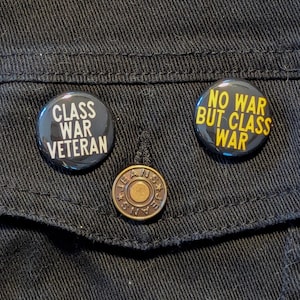 May include: Two black button pins with yellow text on a black denim jacket. The pins say "Class War Veteran" and "No War But Class War".