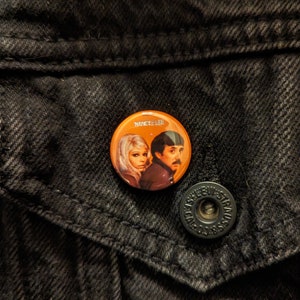 May include: Orange button badge with a picture of Nancy Lee, a woman with blonde hair and a man with brown hair. The text "NANCY LEE" is printed on the badge.