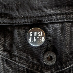 May include: A black and white button with the text "Ghost Hunter" on a black denim jacket.