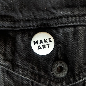 May include: White button with black text that reads "MAKE ART" on a black denim background.