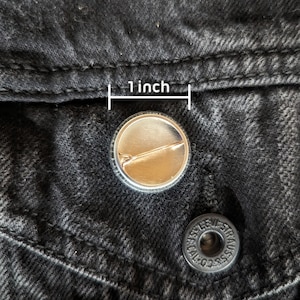 May include: A 1-inch round button pin with a metallic silver finish, set against a black denim jacket. The pin's design is simple, with a shiny surface and a safety pin back. A second button is visible below, with the text "LEVI STRAUSS & CO."