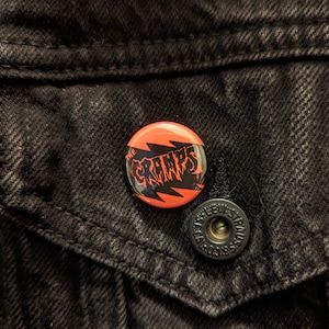 May include: A round button pin with the text "THE CRAMPS" in black, stylized lettering, set against a red and black lightning bolt design. The pin is affixed to a black denim jacket.