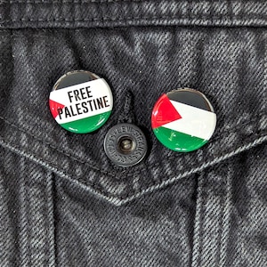 May include: Two round buttons with the Palestinian flag design. One button has the text "FREE PALESTINE" in white. The buttons are on a black denim jacket.