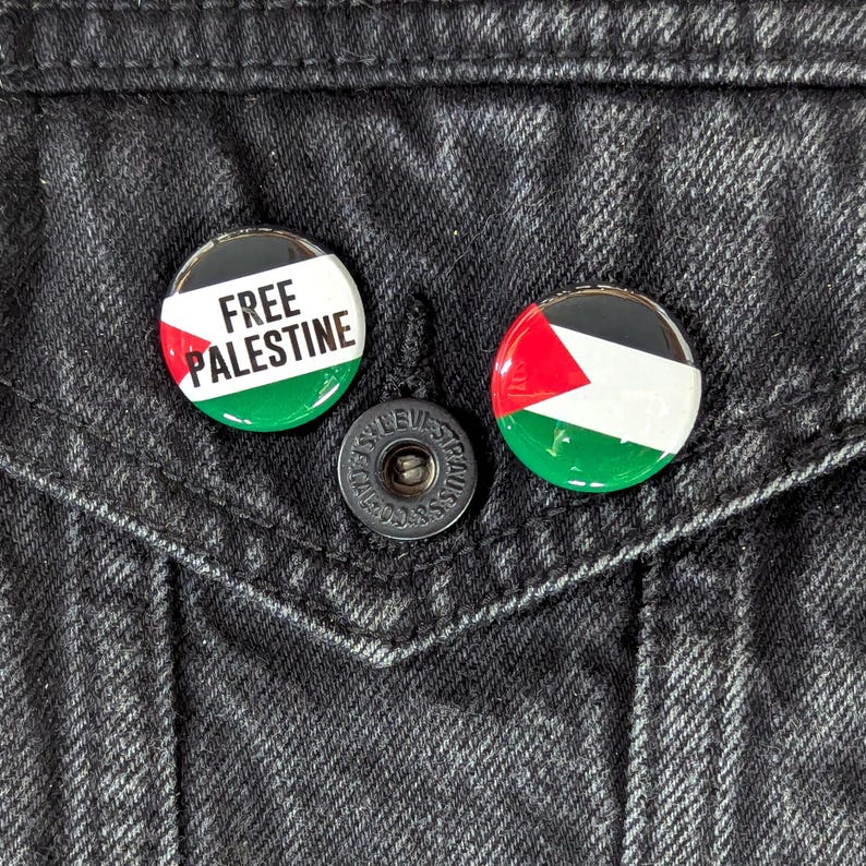 May include: Two round buttons with the Palestinian flag design. One button has the text "FREE PALESTINE" in white. The buttons are on a black denim jacket.