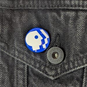 May include: A blue and white PBS logo pin is affixed to a black denim jacket. The pin is round and features the iconic PBS logo. A silver button with the text "LEVI STRAUSS & CO." is also visible.