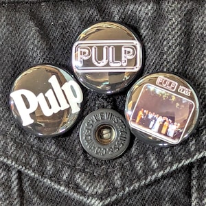 May include: Four round, shiny buttons with the word "Pulp" in white lettering. One button has a photo of a band. A black button with the word "REUSS" is also visible. The buttons are on a black denim background.