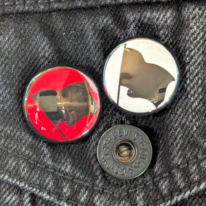 May include: Close-up of three metal buttons on a dark denim fabric. Two buttons are round with graphic designs: a red heart and a black flag. The third button is a smaller, dark gray button with the text "LEVI STRAUSS" around the edge.