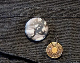 May include: A black denim jacket with a button featuring a black and white photo of a person playing a musical instrument and a gold metal button with the word "JEANS" written on it.
