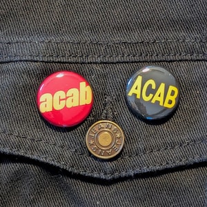 May include: Two black and yellow buttons with the text "ACAB" on a black denim jacket. One button is red with the text "acab" in yellow.