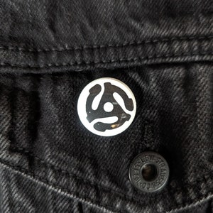 May include: A white button with a black vinyl record symbol on a black denim jacket.