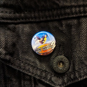 May include: A round button pin featuring the text "Tony Hawk's Pro Skater 2" with an image of a skateboarder in action. The pin has a blue, yellow, and brown color scheme and is set against a black denim jacket.