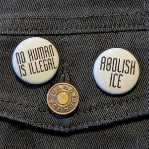 May include: Three white button pins with black text. The pins say "No Human Is Illegal", "Abolish ICE", and "Jeans". The pins are on a black denim jacket.