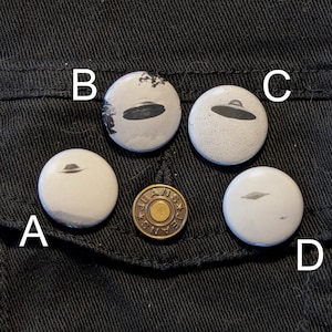 May include: Four white round buttons with black and white images of UFOs on a black denim jacket. The buttons are labeled A, B, C, and D.
