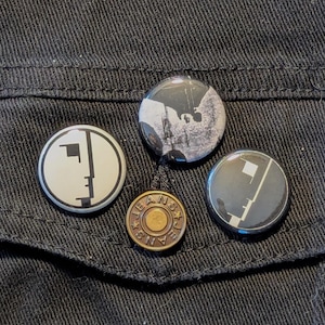May include: Four black and white button pins with different designs. The pins are on a black denim jacket. One pin has a black and white image of a person, one pin has a black and white image of a building, one pin has a black and white image of a face, and one pin has a black and white image of a geometric shape.