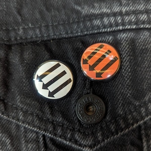 May include: Two black and white button pins with an arrow design. The pins are on a black denim jacket.