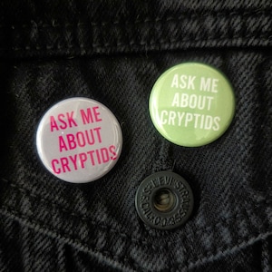May include: Two round buttons with the text "Ask Me About Cryptids" in white letters on a green and a gray background. The buttons are pinned to a black denim jacket.