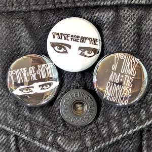 May include: Four round buttons and a black button are displayed on a dark denim surface. Three buttons feature the text "Siouxsie and the Banshees" with eye graphics. One button is white with black text and eyes. The black button has text.