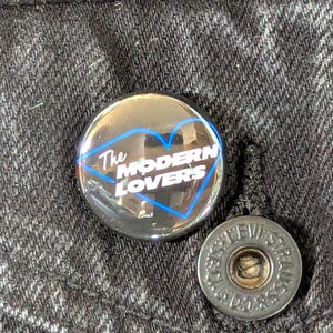 May include: A round button with the text "The Modern Lovers" in white, inside a blue heart-shaped outline. A silver-colored button is next to it, with the text "LEVI STRAUSS & CO." around the edge.