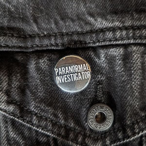 May include: A silver button pin with the text "Paranormal Investigator" in white lettering on a black denim jacket.