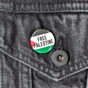 May include: A round button pin with the text "FREE PALESTINE" in white, set against the colors of the Palestinian flag: red, green, and black. The pin is attached to a black denim jacket.
