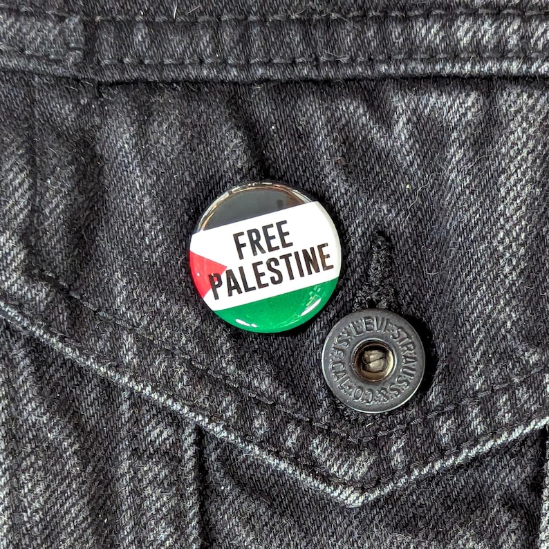 May include: A round button pin with the text "FREE PALESTINE" in white, set against the colors of the Palestinian flag: red, green, and black. The pin is attached to a black denim jacket.