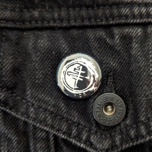 May include: A black and white button with a stylized graphic of a cross inside a circle. The button is on a black denim jacket.