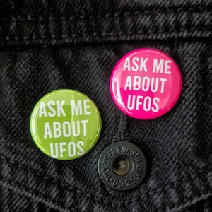 May include: Two round button pins with the text "Ask Me About UFOs" in white letters. The left pin is green and the right pin is pink.