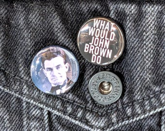 May include: Two circular pinback buttons attached to a dark gray denim fabric. One button features a portrait of a man with dark hair and a bowtie. The other button is black with white text that reads, WHAT WOULD JOHN BROWN DO.