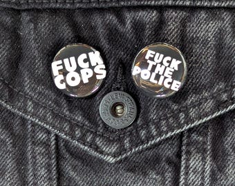 May include: Two round, black and silver pinback buttons with white text that reads "FUCK COPS" and "FUCK THE POLICE" are pinned to a black denim jacket. A silver button is centered below the pins.