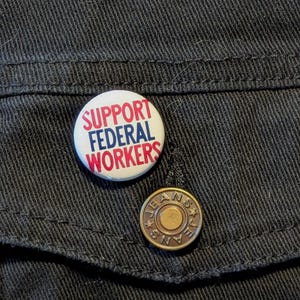 May include: A white button with red and blue lettering that reads "Support Federal Workers" on a black denim jacket.