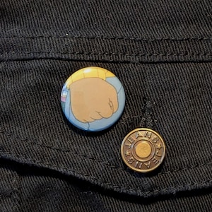 May include: A close-up of two buttons on a black denim jacket. The top button is a round, colorful button with a cartoon image of a fist. The bottom button is a round, gold-colored button with the word "JEANS" embossed on it.