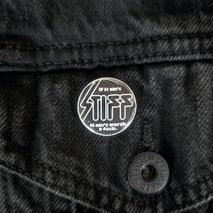 May include: A round, silver-colored pin with the word "STIFF" in bold, stylized letters. The pin also features the text "If it ain't stiff, it ain't worth a fuck." The pin is attached to a black denim jacket.