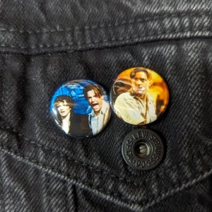 May include: Two round button pins featuring characters from the movie Titanic. The pin on the left features a woman with short brown hair and a man with brown hair. The pin on the right features a man with short brown hair and a blue shirt.