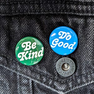 May include: Two circular pinback buttons attached to black denim fabric. One button is green with the white text Be Kind in a cursive font. The other button is blue with the white text Do Good in a cursive font. A metal button snap is visible.